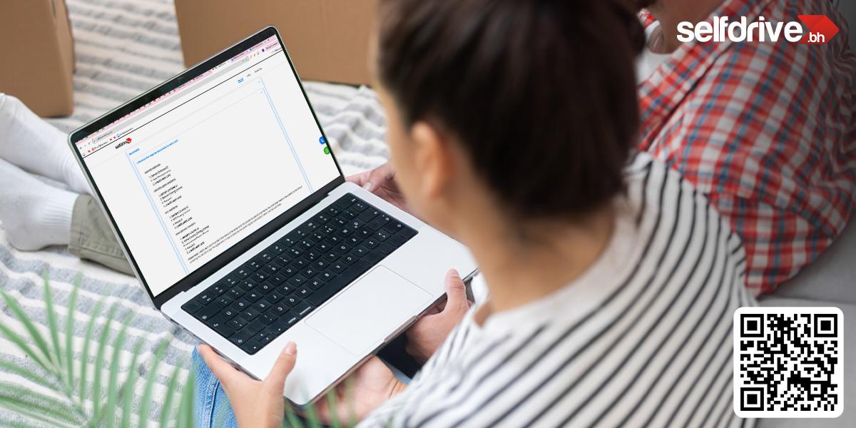 A person is sitting on a bed holding an open laptop, displaying a document. Boxes appear in the background. The scene conveys a relaxed, casual setting.